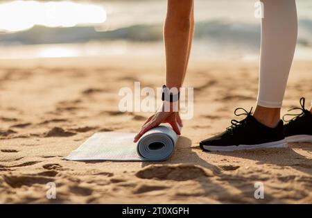 Fit lady putting mat on sand, preparing for training on ocean beach at spare time on sunny day, closeup, side view Stock Photo