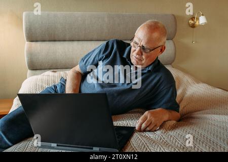 An elderly man is lying on his bed, having fun with his laptop, working remotely, surfing social networks on the Internet. Stock Photo