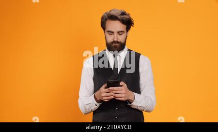 Competent waiter checks smartphone, ready to serve clients at table. Sophisticated, trendy staff member who works in culinary industry depends on mobile phone application, messaging. Stock Photo