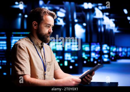 Skilled mechanic resolving performance related issues in server room. Expert utilizing tablet to identify operational problems causing high tech facility electronics slowdown Stock Photo