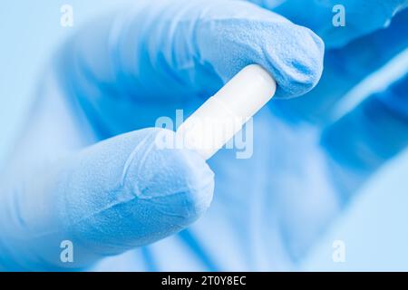 Doctor, pharmacist or scientist hand in glove holding white capsule of pill, vitamin or supplement on blue background. Medicine or drug therapy Stock Photo