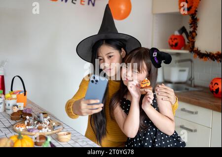 A cute and happy young Asian girl in a Halloween costume is taking selfies with her mom, celebrating Halloween at home together. Stock Photo