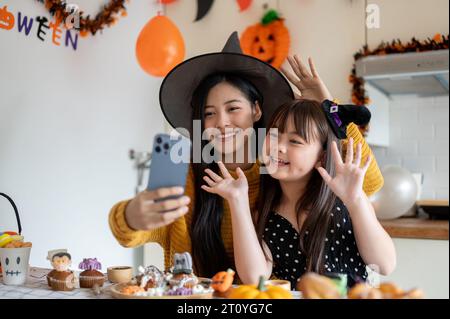 A cute and happy young Asian girl in a Halloween costume is having fun, taking selfies with her mom, and celebrating Halloween at home together. Stock Photo