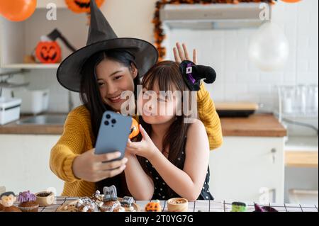 A cute and happy young Asian girl in a Halloween costume is having fun, taking selfies or taking video with her mom while making Halloween cupcakes an Stock Photo