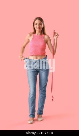 Beautiful young woman in loose jeans with measuring tape on pink background. Weight loss concept Stock Photo