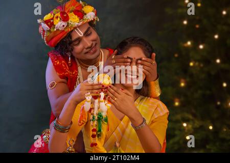 Young man dressed up as Lord Krishna and covering eyes of woman on the occasion of Janmashtami Stock Photo