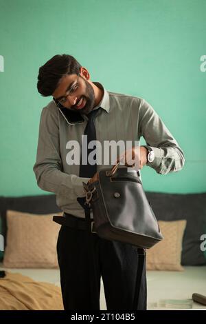 Young man packing his bag and talking on mobile phone while getting ready for office Stock Photo