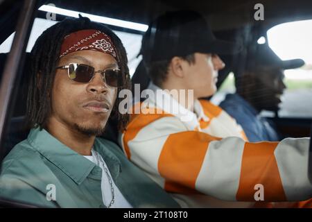Young African American man in sunglasses and casual attire sitting on backseat of in car against two friends and looking through window Stock Photo