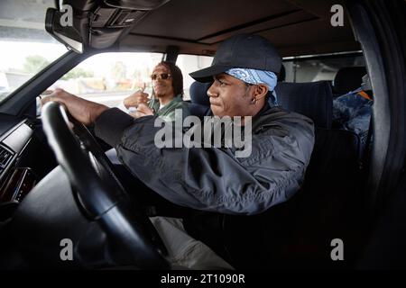 Side view of young hip hop dancer in black bomber jacket and cap driving car while sitting on front seat against his buddy in sunglasses Stock Photo