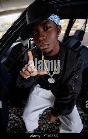 African American hip hop dancer in black bomber jacket, cap and white jeans sitting by open door in front seat of car and looking at camera Stock Photo