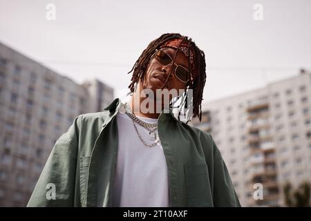 Serious young African American man in sunglasses and stylish casualwear looking at camera while standing against tall buildings outdoors Stock Photo