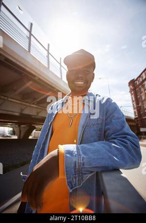 Young smiling African American man in cap and denim jacket standing by railings on bridge and looking at camera against sunlight and blue sky Stock Photo