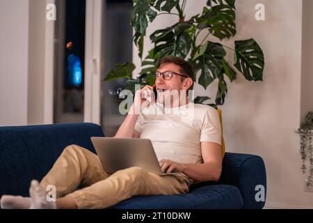 Surprised middle aged man talking on mobile phone lying on couch with laptop Stock Photo