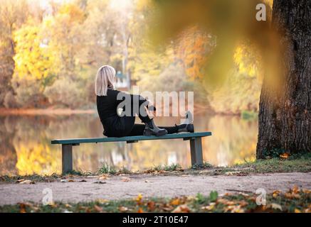 A blonde woman is relaxing on a sunny evening, sitting on a park bench near a pond and admiring the autumn view Stock Photo