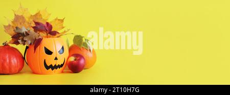 Happy Halloween. Plastic pumpkin bucket with an evil smiling, leaves and ripe pumpkins on a yellow background, empty space for text. Banner for Hallow Stock Photo