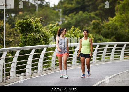 two young asian female joggers talking chatting while walking outdoors in city park Stock Photo
