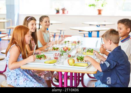 Happy kids having meal with cutlery while sitting together at table in school canteen Stock Photo