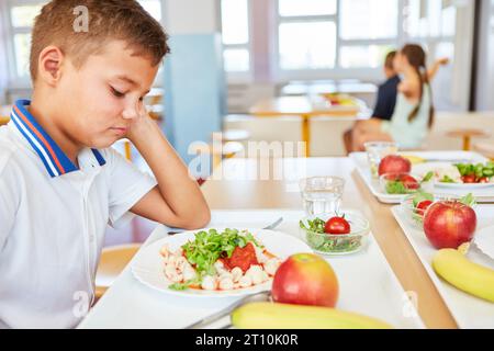 Sad schoolboy looking at food plate while sitting in school cafeteria Stock Photo