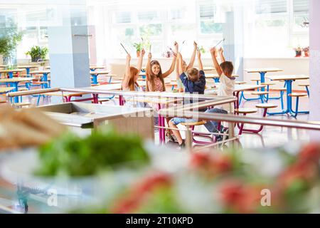 Cheerful male and female students with arms raised sitting together at table during lunch break in school canteen Stock Photo