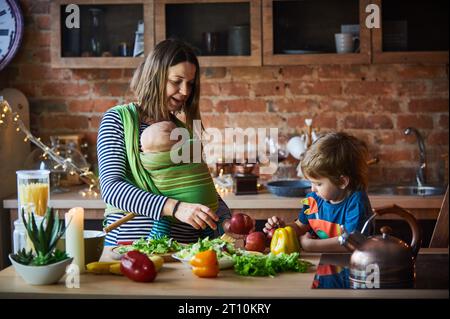 Young family, mother with two children, adorable preschool boy and baby in sling cooking together in a sunny kitchen. Stock Photo