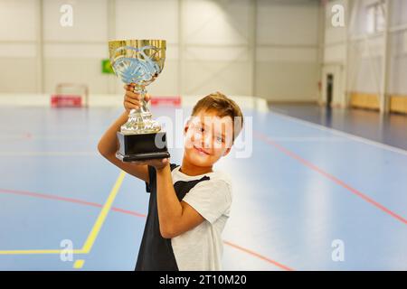 Portrait of smiling schoolboy holding trophy while standing in school gym Stock Photo