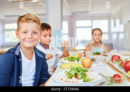 Portrait of smiling blond boy sitting with healthy food plate on table in school cafe Stock Photo
