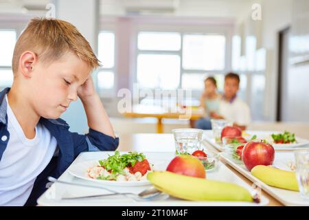 Worried blond boy staring at food plate while sitting at table in school cafeteria Stock Photo