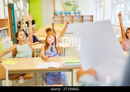 Male and female students raising hands while sitting together during lecture in classroom Stock Photo