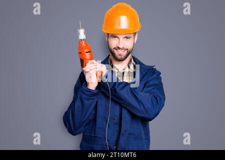 Portrait of joyful positive repairer in protective hard hat with stubble showing, having equipment in arm isolated on grey background Stock Photo