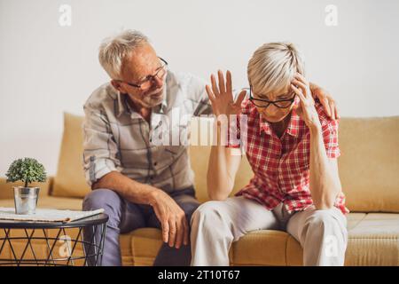 Senior couple sitting in living room and arguing. Woman is angry while man is apologizing. Stock Photo