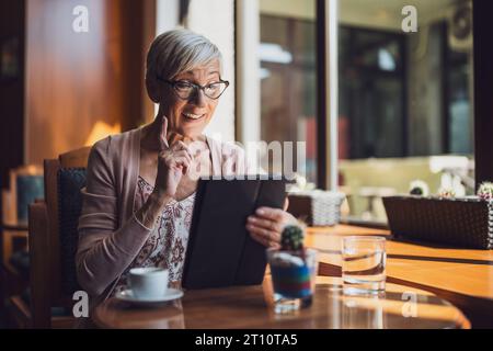 Mature woman is sitting in cafe and relaxing. She is using digital tablet. Stock Photo