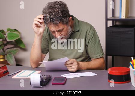 A fifty-year-old man with myopia, adorned with gray beard, lifts his glasses to closely read a letter, possibly a bill. He's casually dressed in a pol Stock Photo