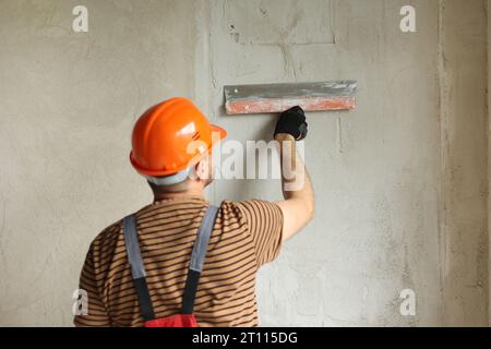 Rear view of handyman worker in overalls and protective helmet plastering concrete wall with putty using a big putty knife. Renovation at apartment an Stock Photo
