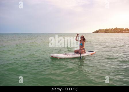 Woman in a blue bikini sits on a sup board with a paddle in the sea against the sunny day Stock Photo