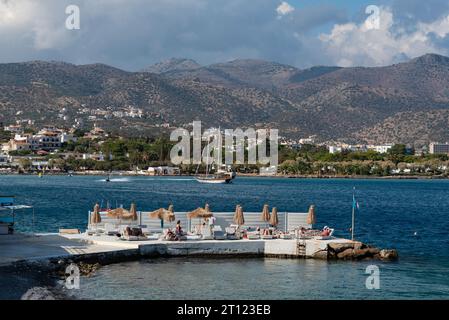 Elounda, Crete, Greece, Europe. 29.09.2023.  Holidaymakers sunbathing on an extended jetty on the Sea of Mirabella and mountains backdrop, Eastern Cre Stock Photo