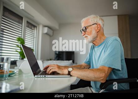 Senior man in wheelchair working from home during retirement. Elderly man using digital technologies, working on laptop. Concept of seniors and Stock Photo