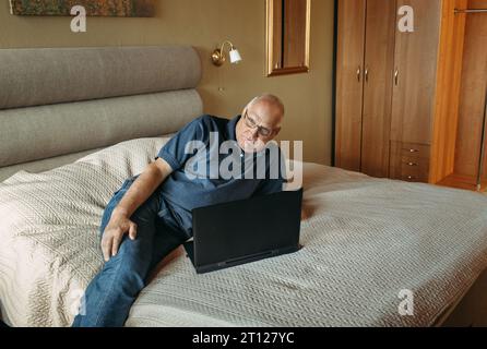 An elderly man is lying on his bed, having fun with his laptop, working remotely, surfing social networks on the Internet. Stock Photo