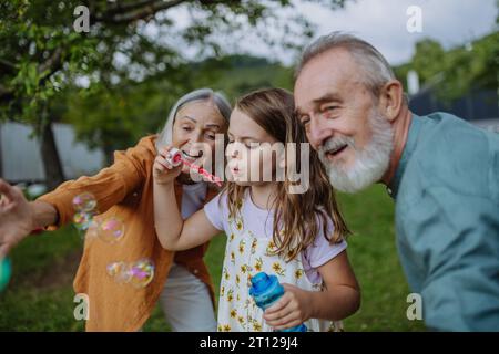 Grandparents and granddaughter are blowing bubbles from a bubble wand, having fun outdoors in the garden during warm autumn day. Stock Photo