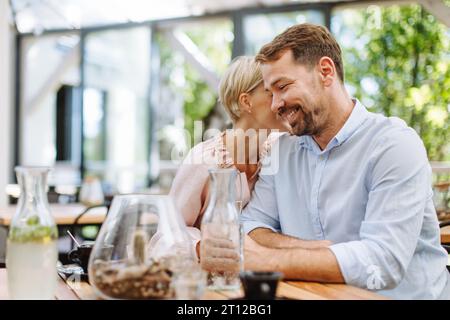 Portrait of beautiful couple in restaurant, on romantic date. Wife whispers into his husband's ear, having romantic moment at restaurant patio. Stock Photo
