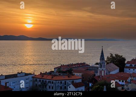 aerial view of the sun setting behind distant mountains in an orange sky reflected in a calm sea from the bell tower of st anastasia in zadar croatia Stock Photo