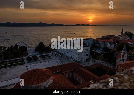aerial view of the sun setting behind distant mountains in an orange sky reflected in a calm sea from the bell tower of st anastasia in zadar croatia Stock Photo