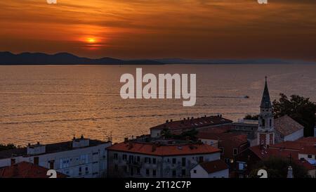 aerial view of the sun setting behind distant mountains in an orange sky reflected in a calm sea from the bell tower of st anastasia in zadar croatia Stock Photo