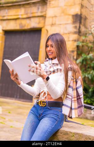 A young Latin woman with a white t-shirt, jeans and a scarf enjoying reading a book, in the old town of Pasaje San Juan, Gipuzkoa, Pais Vasco Stock Photo
