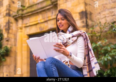 A young Latin woman with a white t-shirt, jeans and a scarf enjoying and imagining reading a book, in the old town of Pasaje San Juan, Gipuzkoa, Pais Stock Photo