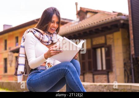 A young Latin woman with a white t-shirt, jeans and a scarf enjoying and imagining reading a book, in the old town. Looking at camera and smiling Stock Photo