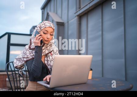 Arab girl with a white veil at the computer on the terrace of a coffee shop, with the computer and talking on the phone Stock Photo