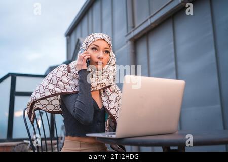 Arab girl with a white veil at the computer on the terrace of a coffee shop, with the computer and talking on the phone Stock Photo