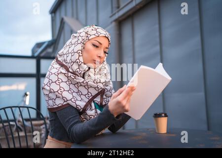 Arab girl with white veil at the computer on the terrace of a coffe shop, having a lot of fun reading a beautiful book Stock Photo