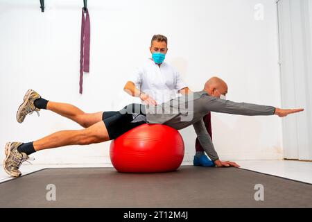 Client balancing on a red ball assisted by Physiotherapist with face mask. Physiotherapy with protective measures for the Coronavirus pandemic Stock Photo