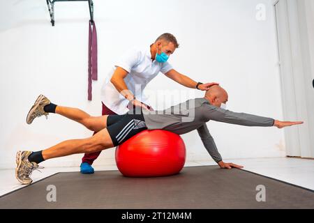 Physiotherapist with face mask helping client to balance on a red ball. Physiotherapy with protective measures for the Coronavirus pandemic Stock Photo